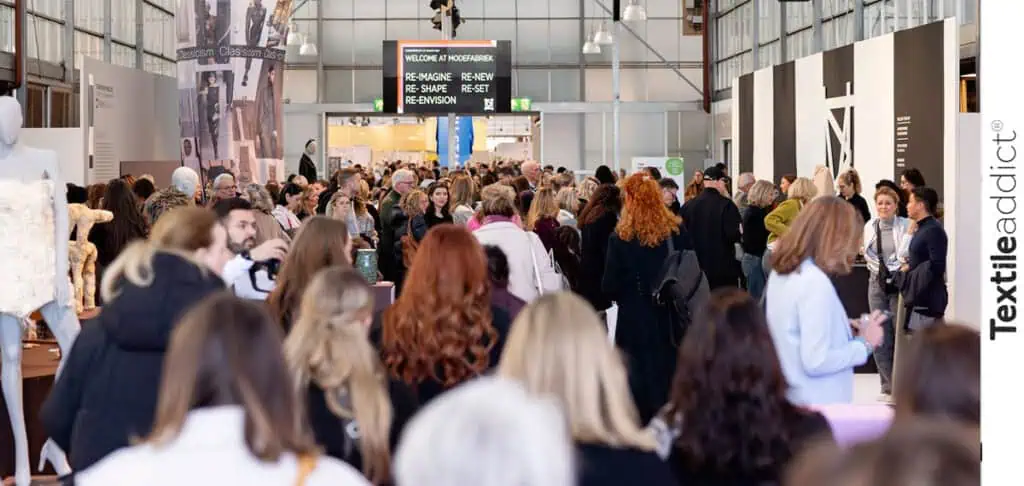 allée des stands animés de Modefabriek Amsterdam, avec des vêtements et accessoires exposés et des professionnels en visite